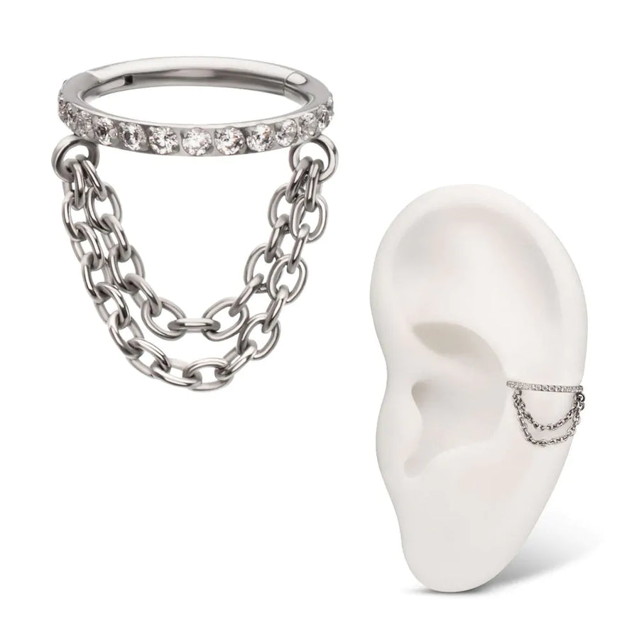 A high-quality silver or titanium hinged clicker jewelry piece featuring a top band set with a row of clear pavé crystals and two draped cable chains of differing lengths hanging below, shown as a standalone item and modeled on a white silicone ear display.