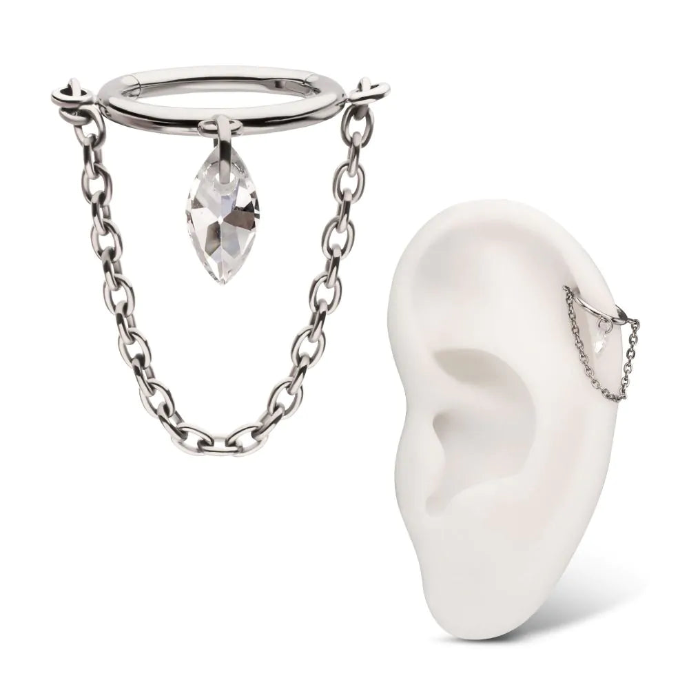A polished silver or titanium hinged clicker jewelry piece featuring a dangling marquise-cut clear crystal and a draped delicate curb chain, shown as a standalone item and modeled on a white silicone ear display.